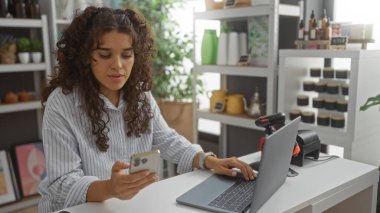 Ev dekorasyonu mağazasında genç bir kadın telefonunu ve laptopunu kullanıyor ve raflarında çeşitli dekoratif eşyalar var.