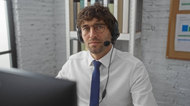 Kulaklık takan adam, modern ofis ortamında bilgisayarın başında arka planda kitap rafları ve çizelgeleri ile güvenle çalışıyor.