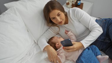 Anne bebek yatağındayken rahat yatak odasında akıllı telefon kullanarak sıcak aile etkileşimini ve modern teknolojiyi gösteriyor..