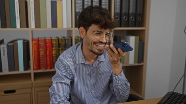 Ofiste telefonda konuşan sakallı yakışıklı genç adam. Arka planda kitap rafları var.