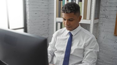 Ofis ortamında bilgisayarda çalışan genç İspanyol bir adam. Resmi giyinmiş, odaklanmış ve profesyonel. Modern bir iç mimarisi var.