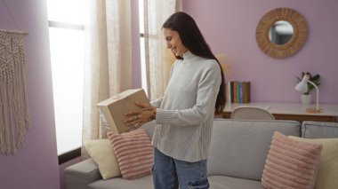 Paketi rahat oturma odasında pembe duvarları olan, makrome duvarlı, bej perdeli, gülümseyen ve kutuya bakan bir kadın..