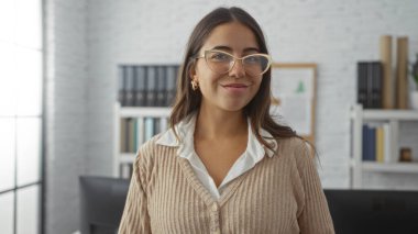 Modern ofis ortamında güvenle gülümseyen, kitaplık takan, gözlük takan, sıradan süveter giyen, profesyonel ama ulaşılabilir atmosferi yakalayan bir kadın..