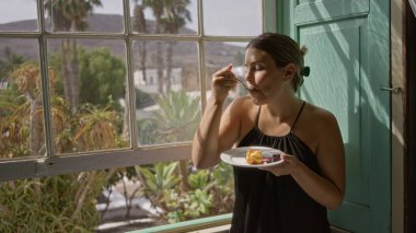 Lokantanın içinde tatlıdan hoşlanan bir kadın açık hava tropikal manzaralı, canlı bir atmosfer ve güneşli bir ortam sergilerken pencerenin yanında duruyor.