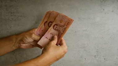 Tayland 'daki finansal kavramları gösteren somut bir arkaplan karşısında Thai baht banknotlarını tutan eller, para birimine yakın çekim odaklı.