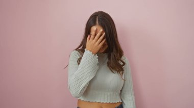 Pembe duvarın önünde eli yüzü kaplayan genç bir kadın, neşeli bir ifadeyle sıradan moda ve güzellik sergiliyor..