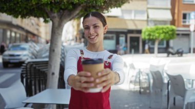 Esmer kadın gülümsüyor ve güneşli bir caddedeki bir kafede kahve ikram ediyor.