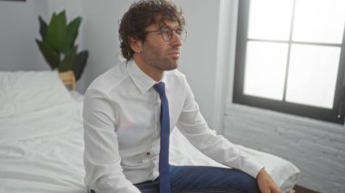 Ofiste gözlük takan genç bir adam penceresi olan bir yatakta düşünceli bir şekilde oturur ve yanında bitki yerleştirir, modern ve yakışıklı bir profesyonel atmosfer taşır..