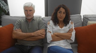 Oturma odasında kolları çapraz oturan orta yaşlı bir çift dekoratif yastıklar ve koltukta yastıklarla çevrili çatışma ve gerilimi dile getiriyorlar.