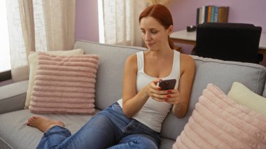 Kanepede uzanan kadın rahat oturma odasında akıllı telefon kullanıyor. Pembe yastıklı. Çekici kızıl saçlı, ev hanımı.