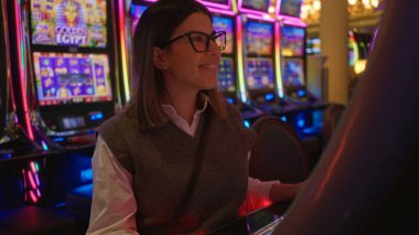 Las Vegas 'ta bir kumarhanede gülümseyen bir kadın. Etrafı renkli kumar makineleriyle çevrili. Canlı, kumar dolu bir ortamda heyecan ve eğlence taşıyor..