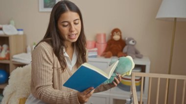 Kadın yatak odasında oyuncak ve beşikle kitap okuyor. İçeride sıcak bir anne havası yaratıyor..