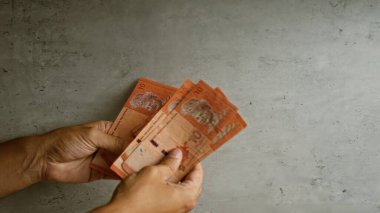 Beton arka planda Malezya halkası tutan adam, erkek eliyle para aktarımını vurguluyor.