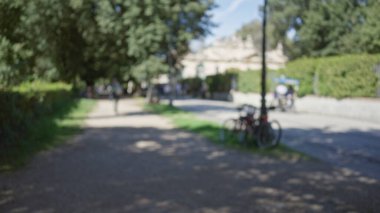 Villa borghese bahçelerindeki bulanık görüntü, Roma 'nın güneşli gökyüzünün altındaki ağaçların, patikaların ve bisikletlerin bulunduğu sakin bir açık hava manzarasını gösteriyor..