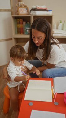 Parlak anaokulunda genç bir çocuğa rehberlik eden bir kadın, neşeli bir ortamda eğlenceli eğitim materyalleriyle çevrili, yetiştirme ile öğretmenliği ve yaratıcılığı vurguluyor..