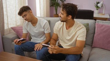 Baba ve oğul evde birlikte vakit geçiriyorlar, kanepede oturuyorlar, akıllı telefonlar kullanıyorlar, rahat bir oturma odasında.