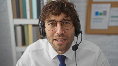 Modern ofis ortamında kulaklık takan İspanyol bir adam bir arkaplan rafa ve ilan panosuna karşı düzenlenen online bir toplantıda odaklanmış ve profesyonel görünüyor..