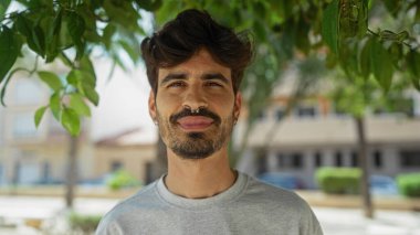 Sakallı yakışıklı İspanyol bir adam. Şehir parkında duruyor. Arka planda binalar ve yeşillikler var.