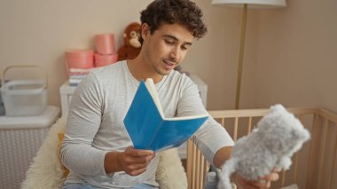 Yanında beşiği olan, yumuşak oyuncaklar ve sıcak bir atmosferle çevrili, rahat bir yatak odasında kitap okuyan genç İspanyol bir adam..