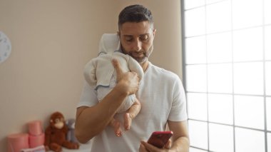Evde bebeği tutan adam parlak oturma odasında akıllı telefona bakıyor yumuşak renklerle huzurlu bir aile anı yaratıyor..