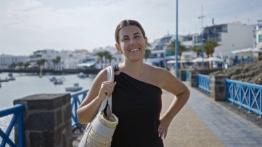 Tutuklu kasabasında, Lanzarote 'de, kanarya adalarında, arka planda güneşli bir günde limanı ve binaları olan açık havada gülümseyen genç bir kadın.