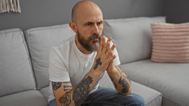 İspanyol dövmeli ve sakallı bir adam oturma odasında bir koltukta oturuyor, derin düşüncelere dalmış, içeride beyaz bir gömlek giyiyor..