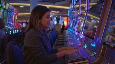 Las Vegas 'ta canlı bir kumarhanede oynayan bir kadın, etrafındaki renkli ışıklarla dikkatle odaklanıyor, kapalı alandaki kumarın cazibesini ve heyecanını yakalıyor..