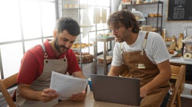 Bir pastanede birlikte çalışan erkekler, ekmek ve hamur işleri raflarıyla çevrili doküman ve dizüstü bilgisayara bakıyorlar, sohbet ediyorlar ve iş operasyonlarını planlıyorlar.