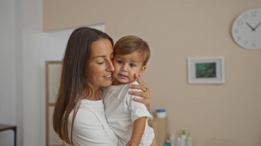 Sıcak bir oturma odasında oğlunu kucaklayan bir kadın aile sevgisini ve İspanyol mirasını sıcak bir tonla evin içinde sergiliyor..