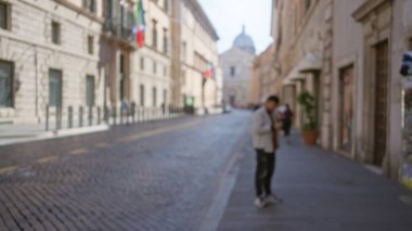 Roma 'da bir kaldırım taşı caddesinde duran bir adam. Bulanık tarihi binalar ve parlak bir gökyüzü altında uzak yayalar..