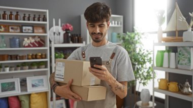 Sakallı yakışıklı genç adam çeşitli dekorasyon malzemeleriyle dolu bir mağazada paketlerini tutarken cep telefonunu kontrol ediyor.