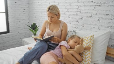 Evladına kitap okuyan bir kadın, rahat bir yatak odasında beyaz tuğladan bir duvarı ve arka planda bir bitkisi olan bir yatakta oturuyor..