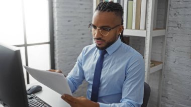 Modern ofis ortamında dokümanları inceleyen, gözlük takan ve resmi kıyafet giyen yakışıklı siyahi bir adam, kapalı bir iş ortamında genç bir profesyoneli örnek alıyor..
