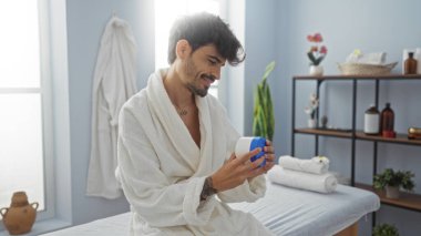 Güzellik ürünlerini elinde tutan yakışıklı İspanyol bir adam spa 'da otururken bornoz giyiyordu. Etrafı kapalı yerlerde sağlık tedavileriyle çevriliydi.