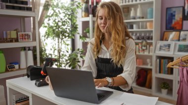 Dekoratörde dizüstü bilgisayarda çalışan bir kadın. Arka planda ev dekoru ürünleri rafları var.