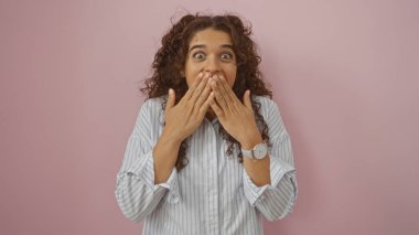 Genç, güzel, esmer, İspanyol bir kadın elleri ağzının üstünde pembe bir arka planda izole edilmiş bir şekilde şaşırdı.