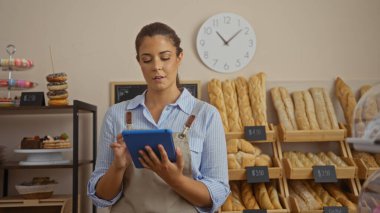 Fırındaki genç kadın tableti kontrol ediyor. Kapalı dükkan ortamındaki ekmekle çevrili.