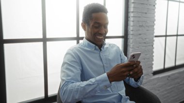 Genç adam iş yerinde kapalı alanda akıllı telefon kullanırken gülümsüyor. Teknoloji odaklı çevreyi gösteriyor.