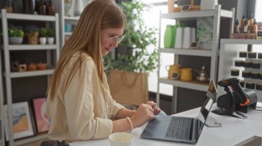 Güzel dekore edilmiş bir mağazada dizüstü bilgisayarda çalışan sarışın bir kadın. Rafları dekorasyon malzemeleriyle dolu.