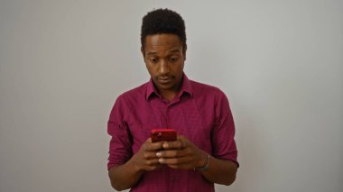 Kırmızı gömlekli genç adam izole edilmiş beyaz arka planda akıllı telefon kullanıyor odaklanmış ve meşgul görünüyor, modern teknoloji etkileşimini vurguluyor.