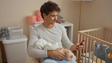 Bebeği kucağında tutan genç adam yatak odasındaki beşiğinden beşiğine akıllı telefon kullanıyor, samimi ve rahat bir ev ortamında modern babalık gösteriyor.