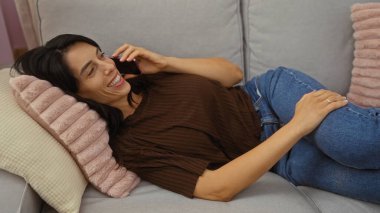 Kanepede uzanmış, rahat bir oturma odasında telefonla konuşan, gülümseyen ve rahatlamış genç bir kadın.