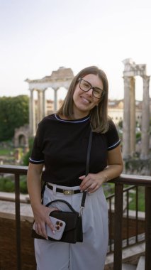 Roma 'da Roma forumunda elinde telefonla gülümseyen, gözlüklü, dışarıda dikilen, tarihi bir Avrupa ortamında İspanyol mirası sergileyen bir kadın..
