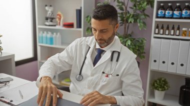 Klinikteki adam beyaz önlük ve steteskop giyiyor, işe odaklanmış, tıbbi malzeme ve belgelerle çevrili, bitkiler ve tıbbi ekipmanlarla donatılmış bir ofiste..