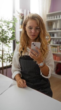 Dekoratörün içinde akıllı telefon kullanan gözlüklü ve önlüklü sarışın bir kadın. Etrafı iç mekan bitkileri ve ev dekorları raflarıyla çevrili..