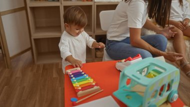Renkli bir anaokulu oyun odasında çocuklarla oturan, ksilofonla oynayan, aile bağlarını ön plana çıkaran bir çocuk..