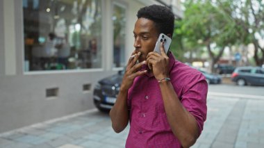 Şehirde sigara içerken akıllı telefon kullanan genç adam gündüzleri açık havada günlük gömlek giyiyor ve ağaçlar ve arabalar görülebilir durumda.