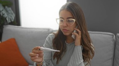 Çağdaş oturma odasında termometre okurken telefonla konuşan bir kadın, ev ortamındaki rahatlık ve teknoloji kullanımını vurgulayarak endişe ve odaklanma gösteriyor..