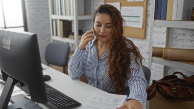 Modern ofiste telefonla konuşan, çizgili gömlek giyen, bilgisayarı, masası, evrakları olan, düzenli çalışma alanı olan, genç, çekici, profesyonel atmosferi olan bir kadın..