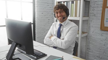 Kulaklıklı yakışıklı adam, ofis ortamında güvenle gülümsüyor, kolları bağlı, modern ev ortamında profesyonelce bir çalışma tutumunu gösteriyor.